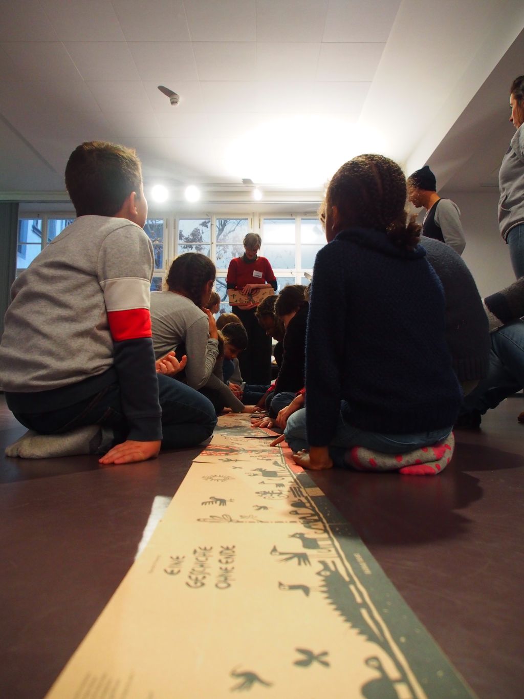 In der Mitte des Bildes liegen ausgelegt die Seiten des Buches, um das sich der Workshop dreht. Die Kinder sitzen entlang der Seiten und lauschen gespannt.